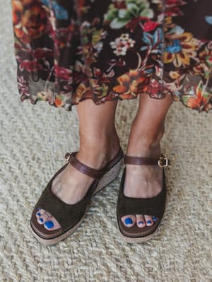 A close-up of olive suede platform wedge sandals with brown leather ankle straps, paired with a richly patterned floral skirt — the kind of shoes that make even a Tuesday feel intentional.