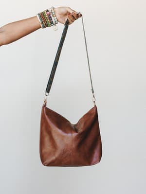 A worn-in caramel shoulder bag held aloft by a beaded-bracelet-stacked wrist — the kind of bag that looks like it's already been somewhere interesting.