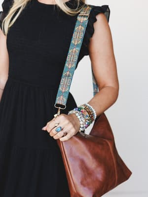 A close-up lifestyle shot of a model in a black ruffle-sleeve tiered dress holding a camel leather crossbody bag with a bold geometric boho-printed strap, her wrist stacked with colorful beaded bracelets and a turquoise stone ring — the kind of styli