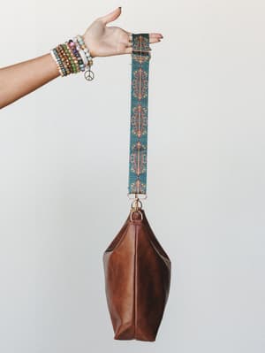 A stacked-bracelet hand holds up a camel leather hobo bag by its wide, kaleidoscopic printed strap — the kind of bag that looks like it already has a story.
