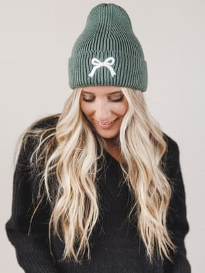 Person wearing a green beanie with a white bow design on a plain background