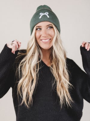 Woman wearing a green beanie with a white logo against a plain background