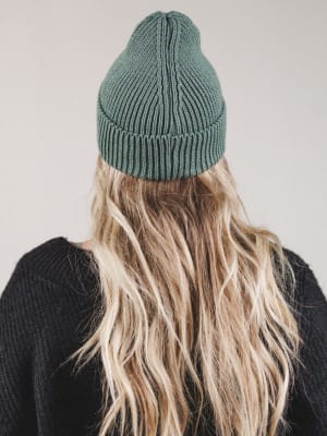 Person wearing a green knit beanie and black sweater with long blonde hair, on a plain background