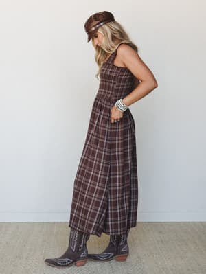 A plaid maxi dress in deep brown gets the full western treatment — smocked bodice, full skirt, cowboy boots, and a leather moto cap stacked with turquoise bracelets for a look that feels like a road trip and a honky-tonk in the same outfit.