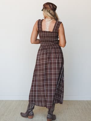 Shot from the back, this brown plaid midi dress has a smocked bodice with lace back detail and a full gathered skirt — styled with cowboy boots and a leather baker boy cap for an earthy, vintage-western feel.