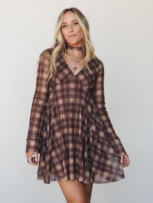 A flowy plaid mini dress in warm brown and cream tones, styled with layered gold necklaces and stacked rings — the kind of dress that feels just right for a fall afternoon that can't decide if it wants to be a concert or a coffee date.