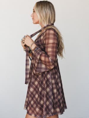 A side-profile shot of a model in a flowy plaid mini dress in warm brown and cream tones, holding the tie neckline detail at her chest — stacked turquoise and gold bracelets and layered rings adding that effortless boho finishing touch.