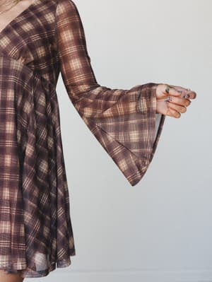 A close-cropped shot of a flowy plaid mini dress in warm brown and cream, with a dramatic bell sleeve and stacked rings on the model's hand giving it that effortlessly vintage, free-spirited feel.