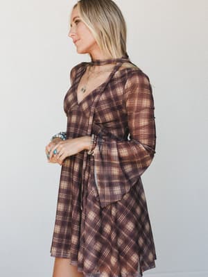 A sheer plaid mini dress in warm brown and cream styled with layered gold necklaces, beaded bracelets, and turquoise rings — the kind of outfit that makes a Tuesday feel like something worth dressing for.