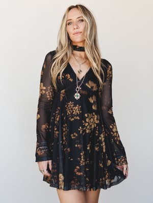 A dark, romantic mini dress in black mesh with golden floral print, styled with layered gold pendants and a velvet choker — the kind of outfit that feels equally at home at a candlelit dinner or a late-summer concert.
