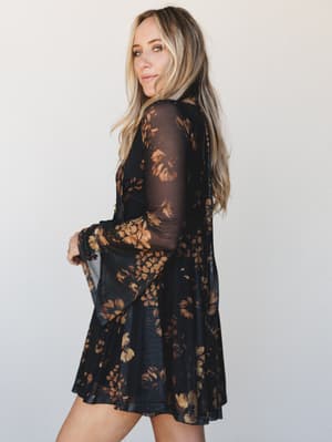 A sheer mesh mini dress in deep black with warm golden floral print — the kind of piece that reads effortless but clearly took thought, shot in profile against a clean studio wall.