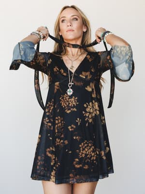 A flowy black mini dress with warm golden-brown floral print and sheer bell sleeves gets a moody, editorial moment — styled with layered gold necklaces and stacked bracelets, the model holds the tie-neck detail up with both hands like she's mid-thoug