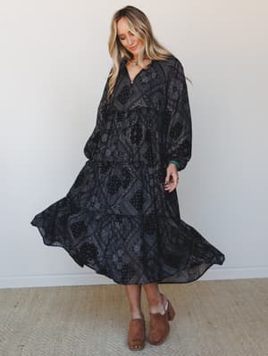 A flowy black maxi dress with an all-over bandana-style medallion print swirls mid-movement, styled with tan wood-platform clogs and turquoise jewelry for a grounded, free-spirited feel.
