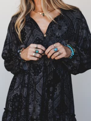 A close-up torso shot of a dark paisley maxi dress styled with layers of boho silver rings, turquoise stones, and beaded bracelets — the kind of outfit that looks like it has a whole story behind it.