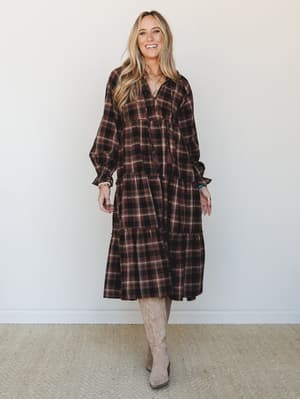 A tiered plaid midi dress in deep brown and cream styled with tall tan cowboy boots and layered boho jewelry — the kind of outfit that feels just as right at a fall farmers market as it does at a weekend dinner.