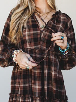 A close-up torso shot of a plaid dress in deep brown and black, styled with layers of beaded bracelets, turquoise rings, and a delicate pendant necklace — the kind of effortless boho jewelry stacking that makes the whole look feel personal and lived-