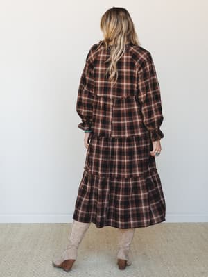 A back-facing shot of a tiered plaid midi dress in deep brown and cream, styled with tan cowboy boots and turquoise jewelry — the kind of outfit that feels equally at home at a fall farmers market or a weekend away.