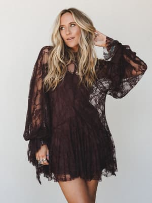 A deep chocolate lace mini dress with billowy long sleeves and a tiered skirt — the kind of dress that feels like a mood, worn by a model with effortless wavy hair and layered rings against a soft neutral studio backdrop.