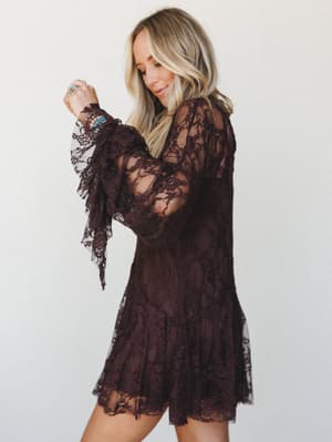 A deep chocolate lace mini dress with sheer bell sleeves and a tiered hem — the kind of piece that feels equally at home at a candlelit dinner or a fall concert, styled here with turquoise stacked bracelets and rings for that signature boho-meets-rom