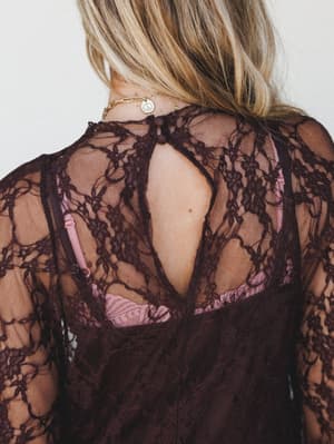 A close-up back detail shot of a deep burgundy lace top or dress, showing a keyhole back closure with a button, floral lace overlay, and a blush pink lining peeking through — the kind of piece that feels intentional from every angle.
