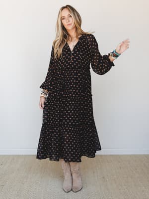 A flowy black midi dress with warm bronze floral dot print, styled with tan suede knee-high boots and stacked bracelets — the kind of outfit that works just as hard at a fall farmers market as it does at dinner.