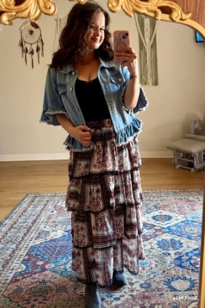A woman takes a mirror selfie indoors wearing the Scottie Ruffle Denim Jacket in light blue over a black top, paired with a long patterned tiered skirt and ankle boots. The room has decorative wall hangings, a patterned rug, and wooden flooring.