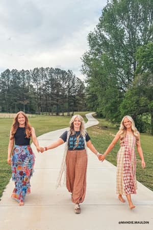 Three women stroll barefoot on a sidewalk, smiling and holding hands. One wears the Thunder Mama Vintage Graphic Tee in charcoal, featuring distressed lettering, while the others don colorful skirts amidst trees and a winding path under cloudy skies.
