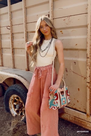 A young woman with long blonde hair stands outdoors by a rusty trailer, wearing a white sleeveless top, layered necklaces, and the Lucky One Wide Leg Pant in mauve, paired with a colorful patterned tote bag on her shoulder.