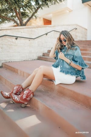 A woman in the Scottie Ruffle Denim Jacket - Light Blue, a white dress, and red cowboy boots sits on outdoor stairs. She has long hair, sunglasses, touches her hair while looking down, with greenery and a building behind her.