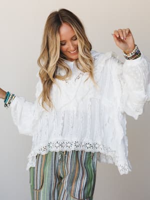 A smiling model dances in a white lace and eyelet ruffle blouse paired with multicolor stripe pants, stacked beaded bracelets on both wrists — the whole look has that joyful, 'spun around in a field' energy.