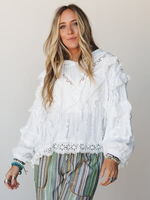 A white lace and ruffle blouse with serious romantic energy, styled with multicolor striped pants and layered beaded bracelets — the kind of outfit that looks like it was effortless but definitely wasn't.