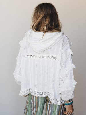 Shot from the back, this white lace-trimmed hooded blouse has a dreamy, handmade quality — ruffled sleeves, eyelet hem detail, and striped boho pants peeking out below, finished with a stack of turquoise and beaded bracelets.