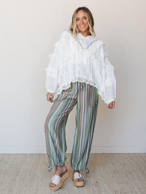 A ruffled white eyelet blouse with lace trim is styled with multicolor striped wide-leg pants and espadrille platform sandals — the kind of outfit that looks like it got dressed for a farmers market and ended up being the most interesting person ther