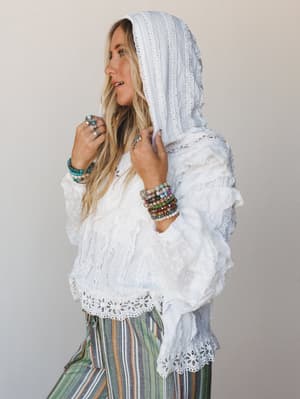 A dreamy white eyelet hooded blouse with ruffled bell sleeves, styled with multicolor striped wide-leg pants and stacked beaded bracelets — the kind of outfit that looks like it was packed for a coastal road trip and never came back.