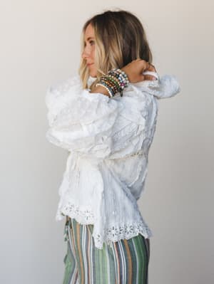A model shot in profile against a soft neutral wall, wearing a billowy white eyelet and ruffle blouse with stacked colorful beaded bracelets and a peek of striped wide-leg pants — the kind of outfit that looks effortless but clearly wasn't an acciden
