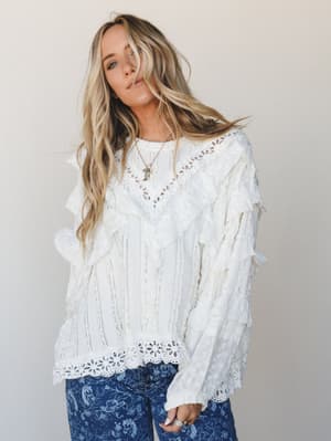 A flowy off-white lace and ruffle blouse with Victorian-meets-boho energy, styled with printed denim and delicate layered necklaces against a soft neutral studio backdrop.