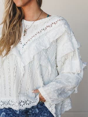 A dreamy off-white blouse layered with eyelet trim and cascading ruffles, styled with a blue floral bottom and delicate gold jewelry — the kind of top that makes a Tuesday feel like something.
