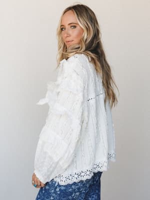 A side-profile shot of a model in an off-white lace and eyelet blouse with ruffled sleeve detailing, paired with a blue floral skirt — the whole look has that effortless, romantic energy of someone who got dressed without trying too hard.