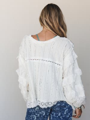 A back-view shot of a flowy off-white blouse with tiered ruffle sleeves, eyelet hem trim, and mixed lace texture paneling — styled with floral print blue jeans and a peek of turquoise bralette, giving it that effortlessly collected, boho-everyday fee