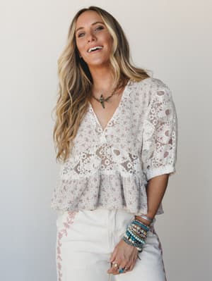 A smiling model in a dreamy mixed-media blouse — ditsy floral print panels meet sheer floral lace inserts — layered with stacked boho bracelets and a bird pendant necklace for that effortlessly personal, every-occasion kind of energy.
