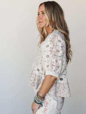 A side-profile shot of a model in a white lace overlay blouse with a ditsy floral underlayer and ruffled hem, paired with white embroidered jeans and a stack of mixed boho bracelets — soft, romantic, and effortlessly lived-in.