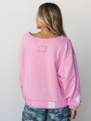 A back-view shot of a bubblegum pink oversized sweatshirt with a peace sign patch at the center back, raw-hem detailing at the hem, lace patches, and camo pants peeking out below — the kind of top that feels intentional without trying too hard.