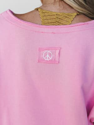 A close-up back detail shot of a candy pink sweatshirt featuring an embroidered peace sign patch at center back, with a peek of a golden lace bralette visible at the neckline.