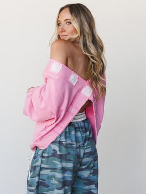 A model poses with her back to the camera, glancing over her shoulder, wearing an off-shoulder candy pink sweatshirt with white lace patch details and a small peace sign emblem, paired with blue-toned camo joggers — effortlessly playful and lived-in.