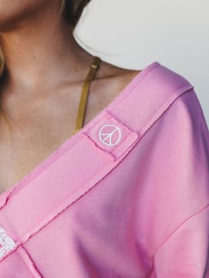 A close-up shoulder detail shot of a candy pink sweatshirt, highlighting a small embroidered peace sign patch and contrast strap trim — the kind of piece that feels personal before you've even put it on.
