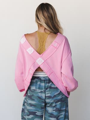 Shot from behind, this candy pink sweatshirt steals the scene with its open cross-back design, lace patches, and peace sign embroidery — styled over a mustard lace bralette and blue camo wide-legs for an effortlessly eclectic, comfort-first look.