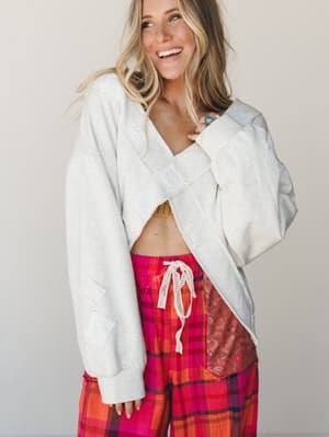 A smiling model wears an oversized cropped oatmeal sweatshirt with patchwork detailing, open at the front and paired with bold pink and orange plaid wide-leg pants — the kind of effortlessly mismatched outfit that somehow just works.