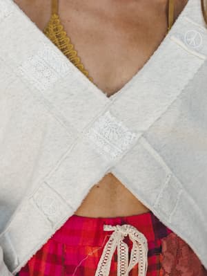 A close-up detail shot capturing the criss-cross front of a cream sweatshirt with lace patch accents and embossed peace sign buttons, layered over a mustard lace bralette and a bold red plaid skirt with a crochet tie waist.