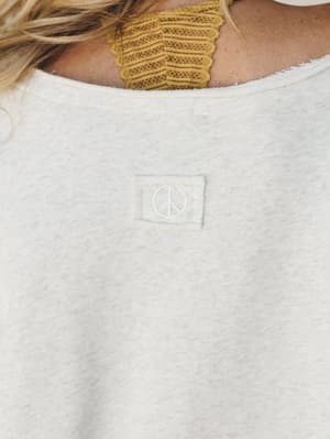 A close-up detail shot of a heathered oatmeal sweatshirt, showing a tonal embroidered peace sign patch at the back neckline — effortlessly boho and worn-in, layered over a mustard lace bralette peeking out at the neckline.