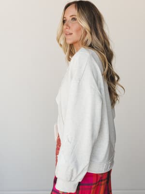 A side-profile shot of a model in a heathered oatmeal pullover sweatshirt with subtle distressed detailing, paired with a bold red and pink plaid skirt — cozy-meets-intentional, the kind of outfit that makes staying in feel like a choice.